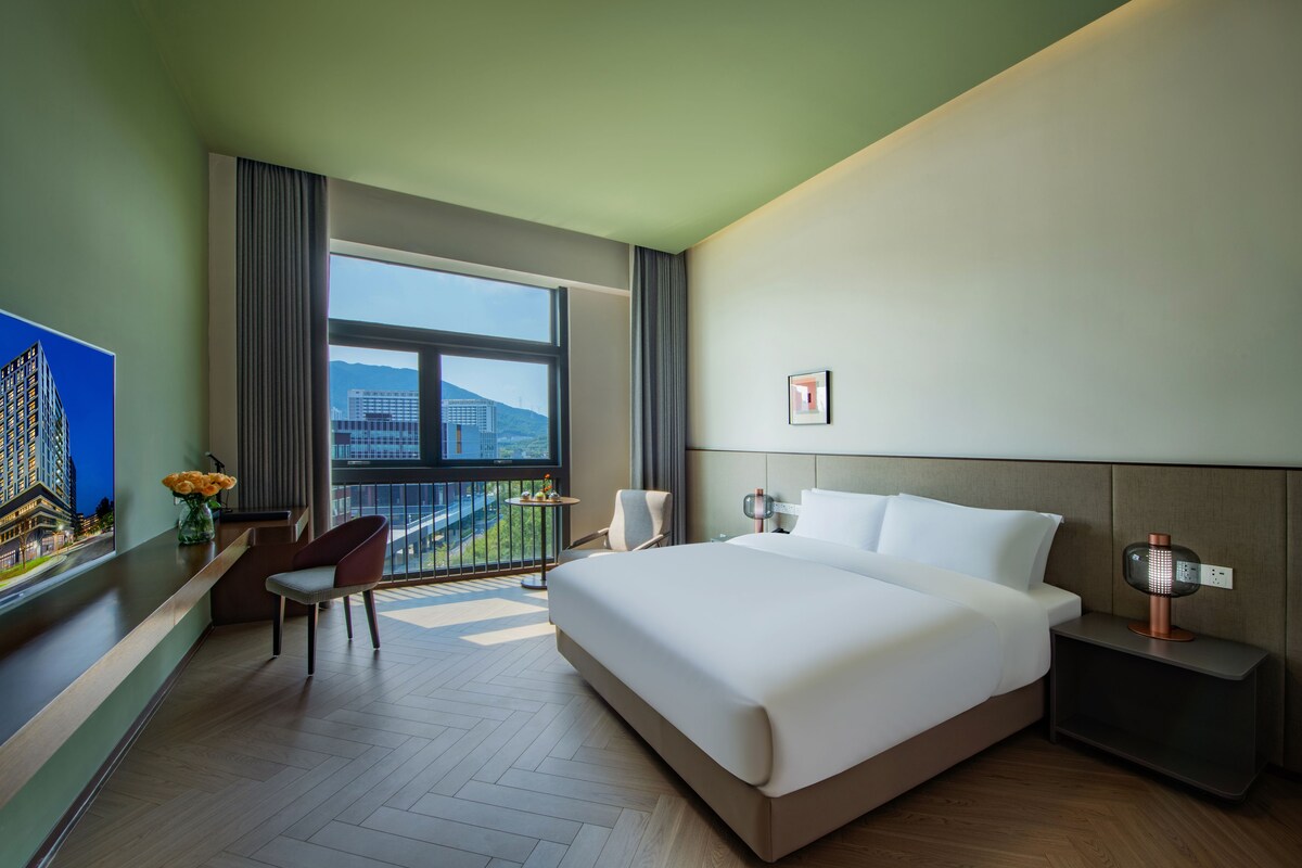 Quarto de hotel com uma cama grande, uma escrivaninha com cadeira, uma mesinha de centro com flores, uma mesinha de cabeceira com abajur e vista para edifícios através de uma janela ampla.