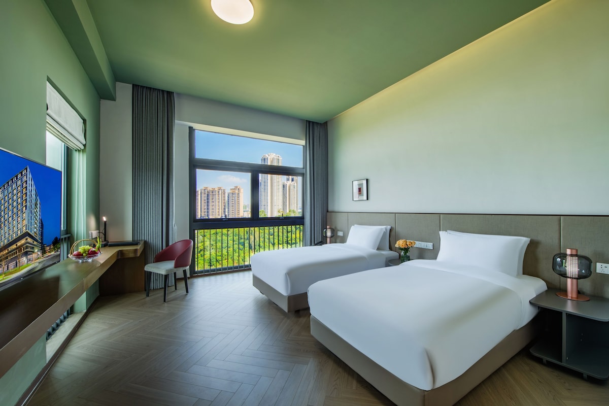 A hotel room with two beds, a desk, a chair, a large window with a view of buildings, and a small table with a lamp.
