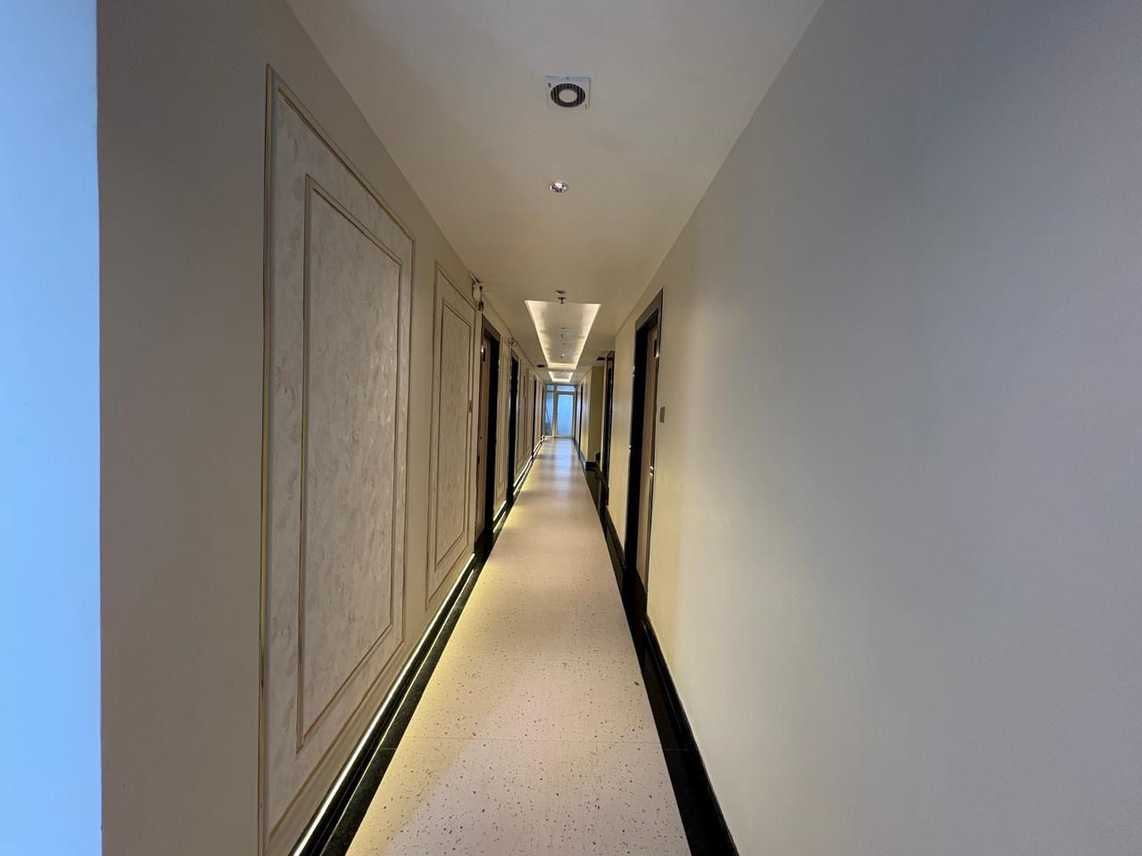 Un long couloir étroit, typique des hôtels, avec des portes des deux côtés, un sol carrelé et un plafond doté d’un éclairage encastré.
