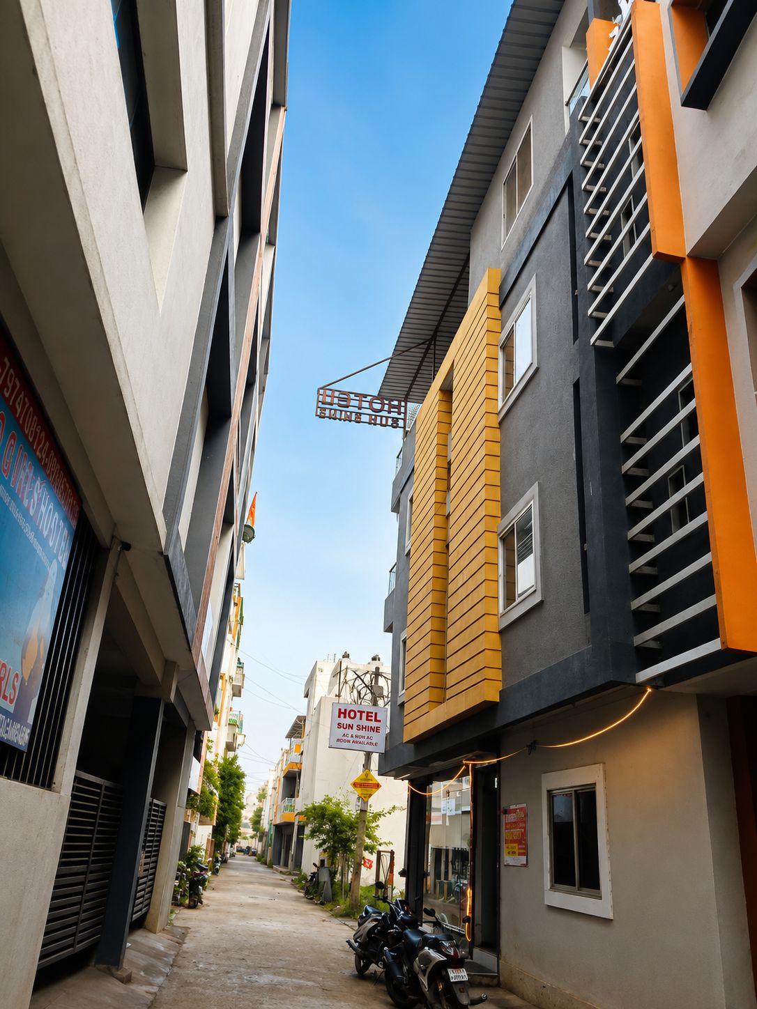 Una calle estrecha con un hotel llamado 'Sun Shine' a la derecha, que presenta una fachada moderna en colores amarillo y gris.