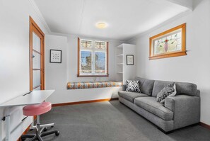 Ein Zimmer mit einem grauen Sofa, einem weißen Schreibtisch mit einem pinken Stuhl und einem Fenster mit Jalousien.
