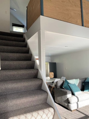 Una escalera conduce a un espacio tipo loft con diseño moderno, un sofá con cojines y una zona de comedor con mesa y sillas.