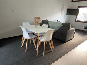 Un coin repas avec une table et des chaises blanches, un canapé gris et un téléviseur fixé au mur.