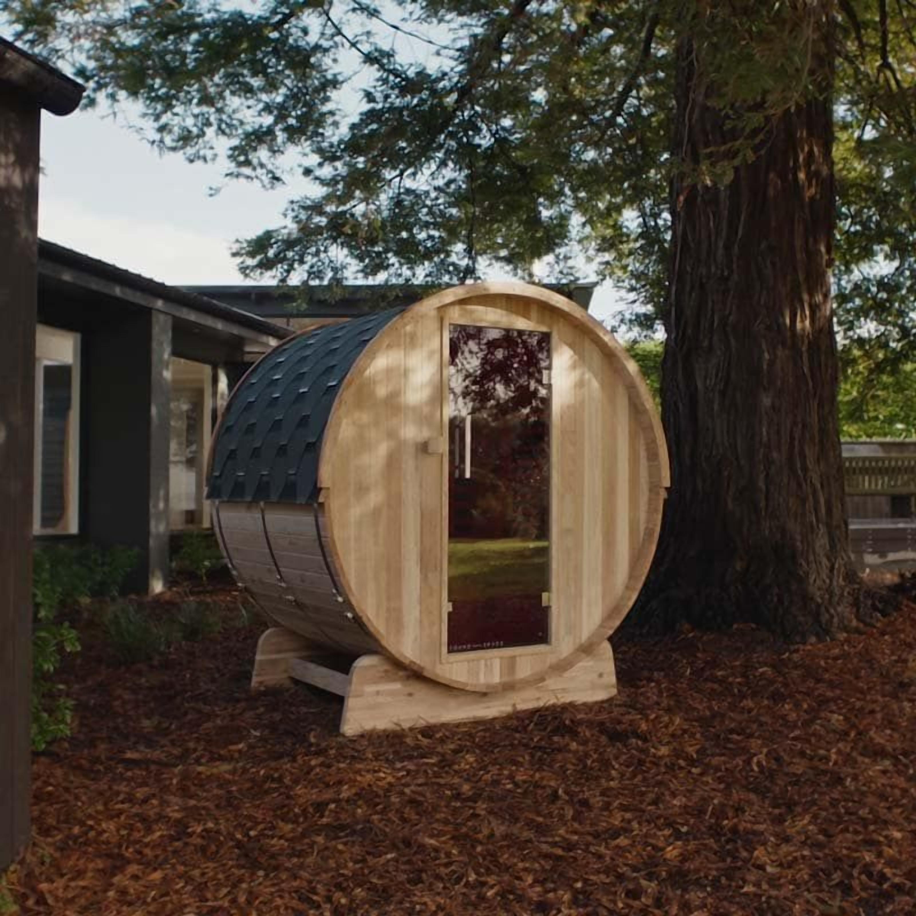 A wooden barrel-shaped structure with a glass door, situated outdoors with a tree and a building in the background.