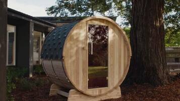 A wooden barrel-shaped structure with a glass door, situated outdoors with a tree and a building in the background.