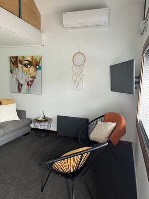 Un salon moderne avec un canapé gris, un fauteuil noir agrémenté d’un coussin orange, une petite table ronde, un attrape-rêves et un téléviseur à écran plat fixé au mur.