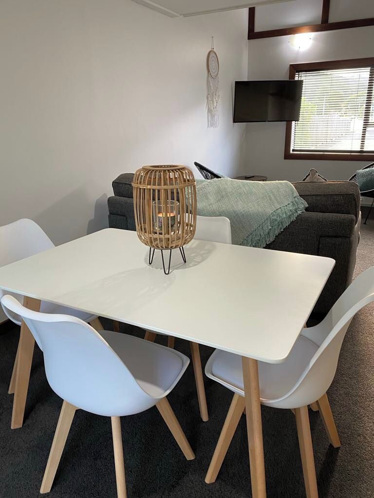 A dining area with a white table and four chairs, a woven light fixture, a grey sofa, and a television mounted on the wall.