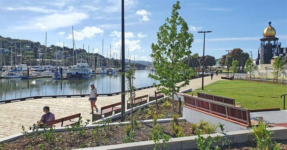 Un port de plaisance avec des bateaux, une promenade piétonne et un espace vert aménagé avec des bancs et des arbres.