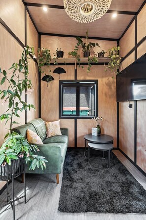 Um quarto aconchegante com um sofá verde, uma mesa redonda pequena e um tapete preto. Há plantas em prateleiras e uma janela com vista.