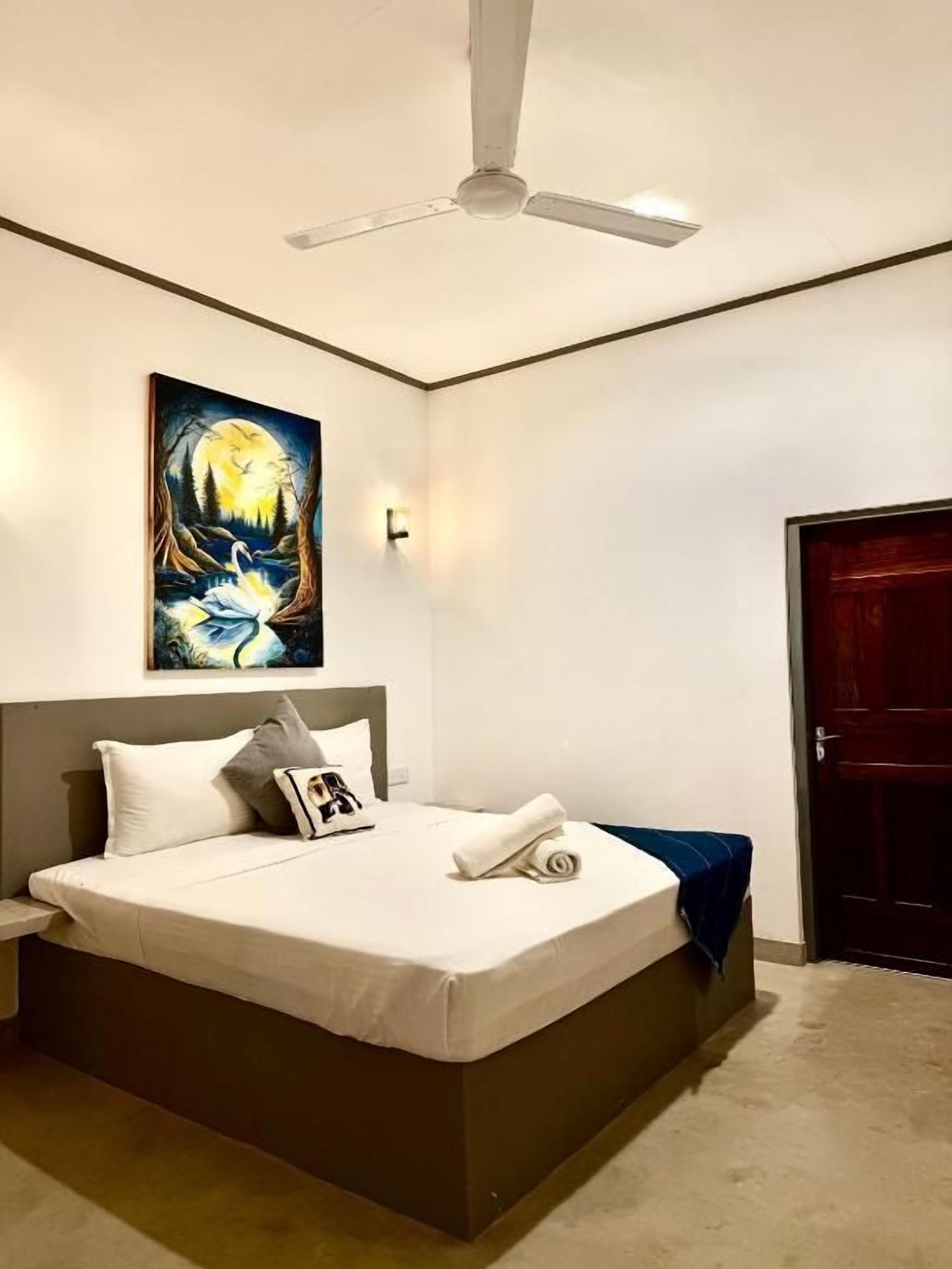 A bedroom with a bed, a ceiling fan, a painting, and a door.