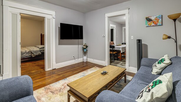 Ein Wohnzimmer mit Flachbildfernseher an der Wand, einem Holztisch und einer Couch mit Zierkissen.