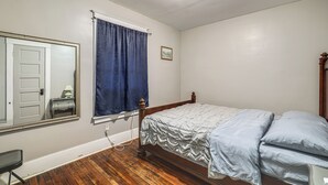 Ein Schlafzimmer mit einem Einzelbett, einem Holzkopfteil, einem Fenster mit blauer Vorhang, einem Nachttisch mit Lampe und einem Spiegel an der Wand.