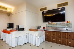 A buffet setup with a flat-screen TV above, a microwave, toaster, and a coffee machine on the counter, and a buffet table with white tablecloths and a red runner.