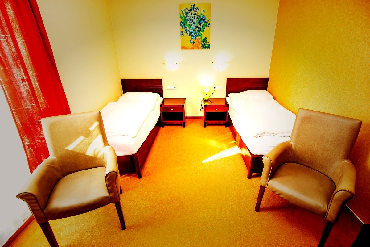 Hotellihuone, jossa on kaksi erillistä sänkyä, yöpöytä lampun kanssa, kaksi tuolia ja taulu seinällä.