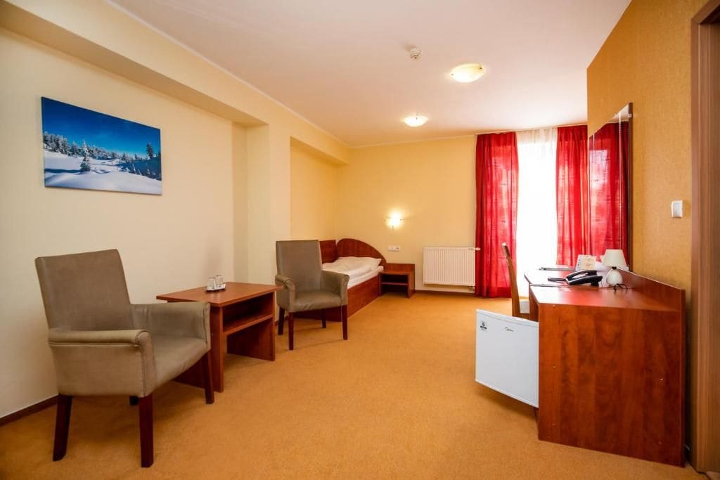 A hotel room with a bed, two chairs, a desk, a radiator, and a painting on the wall.