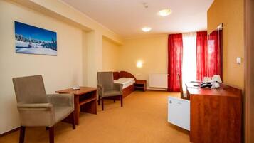 A hotel room with a bed, two chairs, a desk, a radiator, and a painting on the wall.