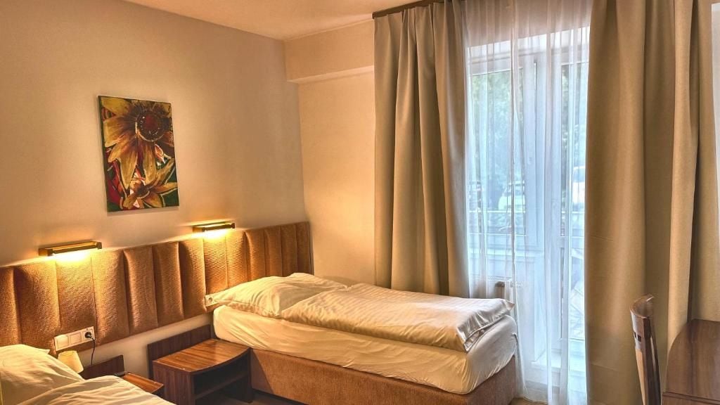 A hotel room with a bed, bedside tables, a painting on the wall, and a window with curtains.