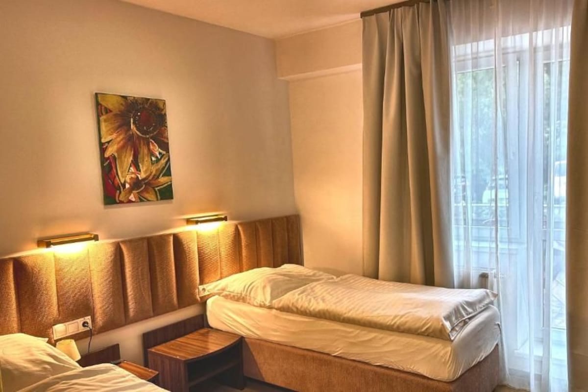 Una habitación de hotel con una cama, mesitas de noche, un cuadro en la pared y una ventana con cortinas.