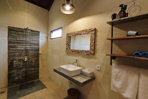 A bathroom with a stone shower, a sink with a mirror, and shelves with towels. - The Daun Bali Canggu (Badung)