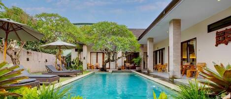 A swimming pool with lounge chairs and umbrellas, surrounded by greenery and a building with a tiled roof.