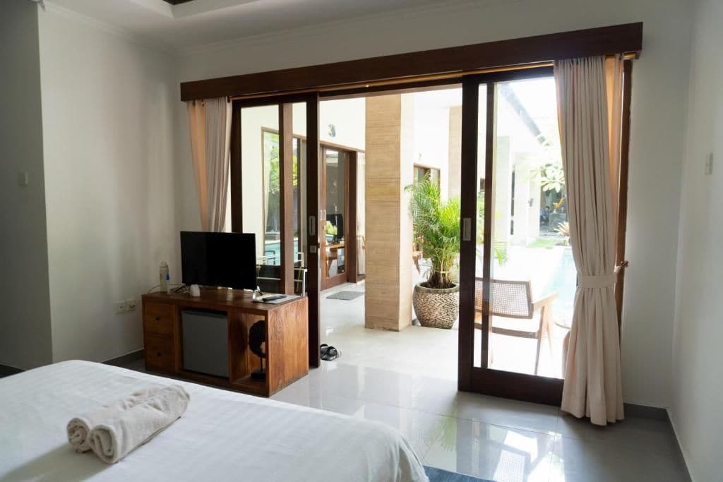A hotel room with a bed, a desk with a television, a potted plant, and a view of an outdoor area with a pool.