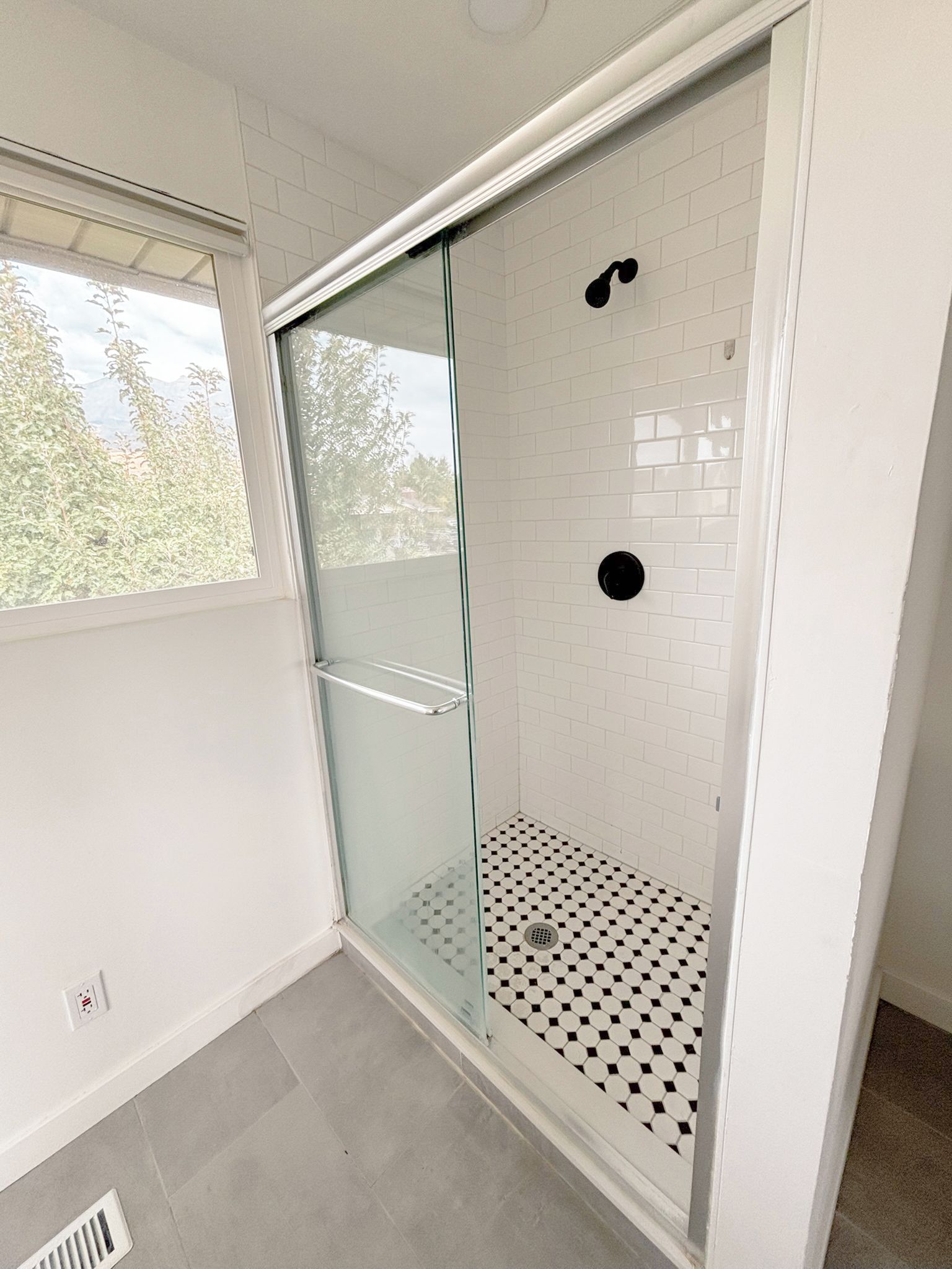 Ein modernes Badezimmer mit einer Duschkabine aus Glas, weißen Fliesen und einem Fenster mit Blick auf Bäume.