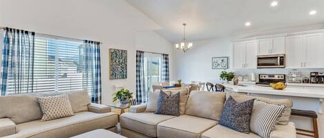 A modern living room with a sectional sofa, a dining area, and a kitchen with white cabinets and a microwave.