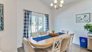 Et spiseområde med et rundt bord, stole, en lysekrone, et vindue med gardiner og en genbrugsbeholder.
