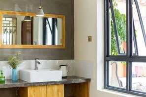 Un baño con un espejo grande, un lavamanos blanco, un tocador de madera y una ventana con vistas al verdor.