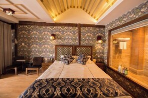 Quarto de hotel com uma cama grande, cabeceira de madeira, escrivaninha e janela com vista.