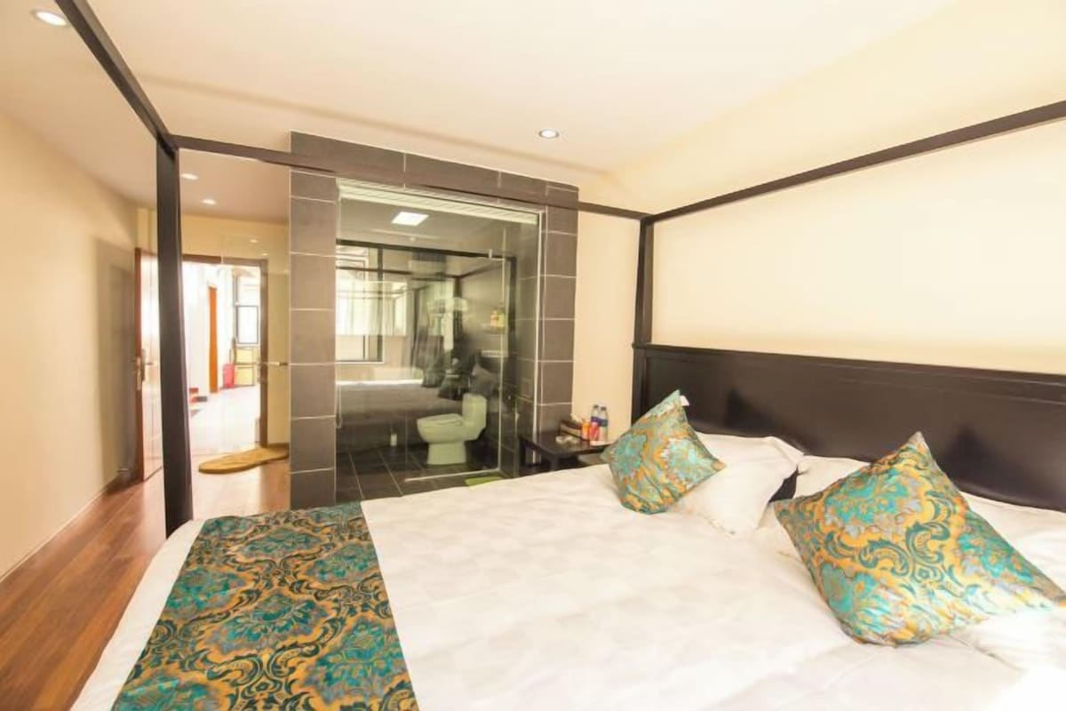 Habitación de hotel con cama, mesita de noche y baño con paredes de vidrio.