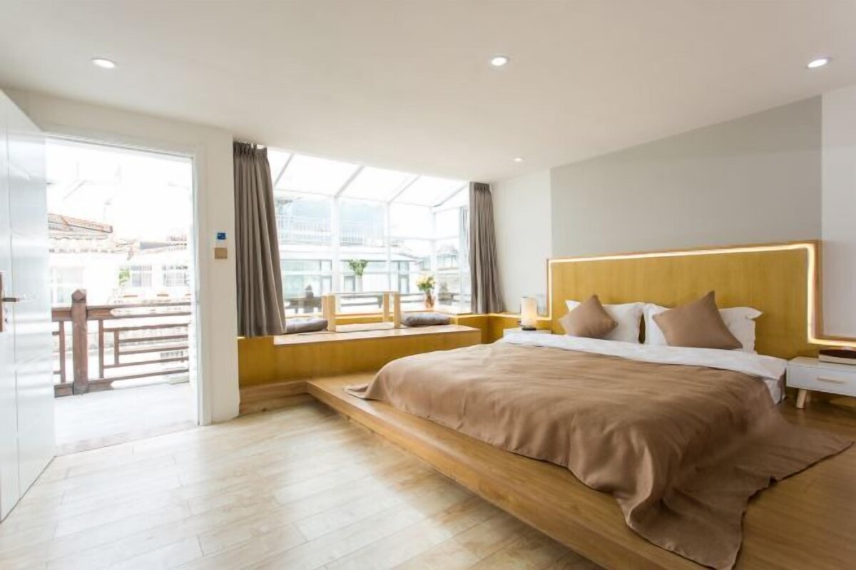 Una habitación de hotel moderna con una cama grande, un cabecero de madera y vistas a la ciudad a través de amplias ventanas.