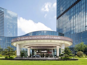A modern hotel with a curved entrance, surrounded by lush greenery and tall buildings.