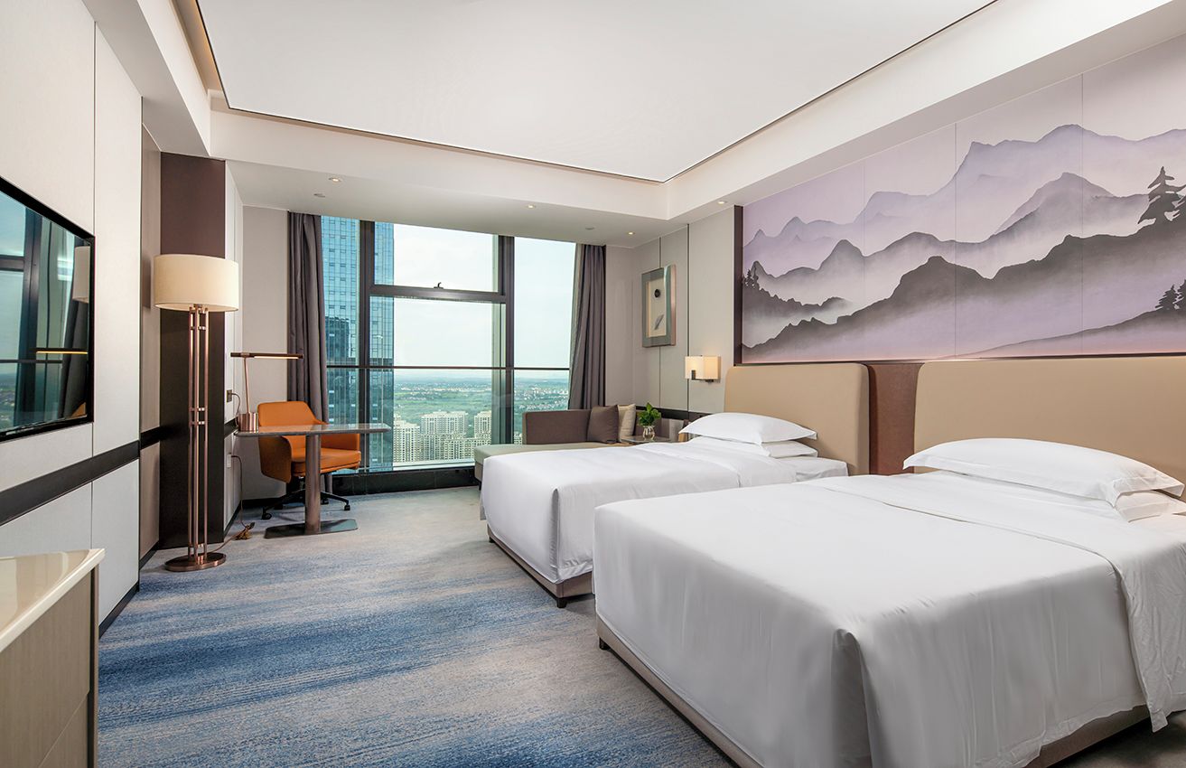 A hotel room with two beds, a desk, a chair, a large window with a city view, and a painting on the wall.