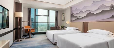 A hotel room with two beds, a desk, a chair, a large window with a city view, and a painting on the wall.