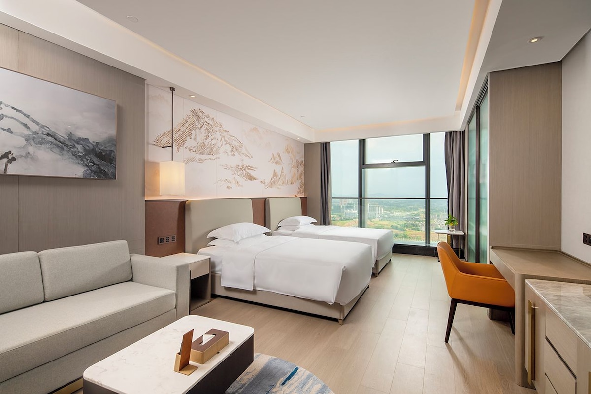 Et moderne hotelværelse med sofa, to senge, et skrivebord og et stort vindue med udsigt over byen.