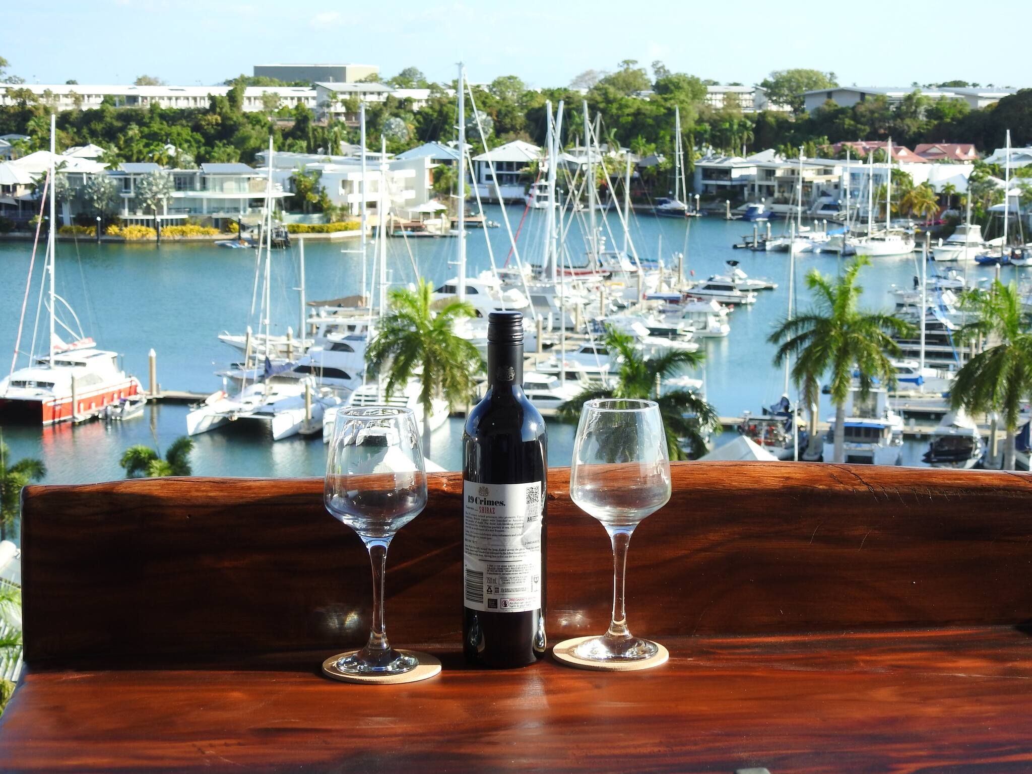Une bouteille de vin et deux verres sur une table en bois, avec une vue sur une marina en arrière-plan.
