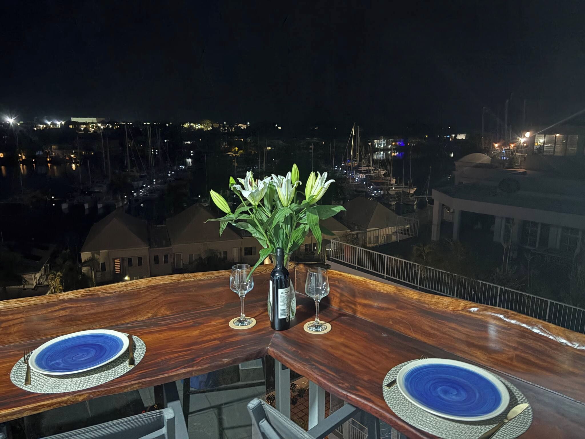 A table set for two with a bottle of wine and glasses, overlooking a marina at night.