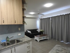 A modern kitchen with a sink, cabinets, and a countertop. There is a black sofa and a coffee table with a vase of flowers. - Luxury Living in Cullen Bay (Darwin City)