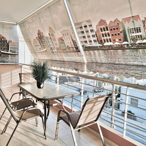 Een balkon met een tafel en stoelen, met uitzicht op een rivier en gebouwen op de achtergrond.
