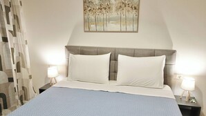En pænt redt seng med knaldet sengetæppe, to hvide puder og en sengegavl. To natborde med lamper står ved sengen, og et maleri hænger over sengen.