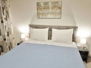 Een net opgemaakt bed met een bedsprei met patroon, twee witte kussens en een hoofdbord. Er staan twee nachtkastjes met lampen en erboven hangt een schilderij.