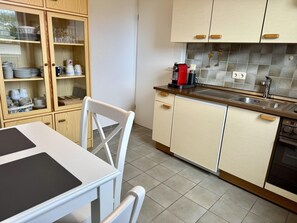 A kitchen with wooden cabinets, a sink, and a dining area with a table and chairs. - SyltRuhe - 2-room apartment for 4 people with a balcony, parking space, and Wi-Fi. Pet (Sylt)