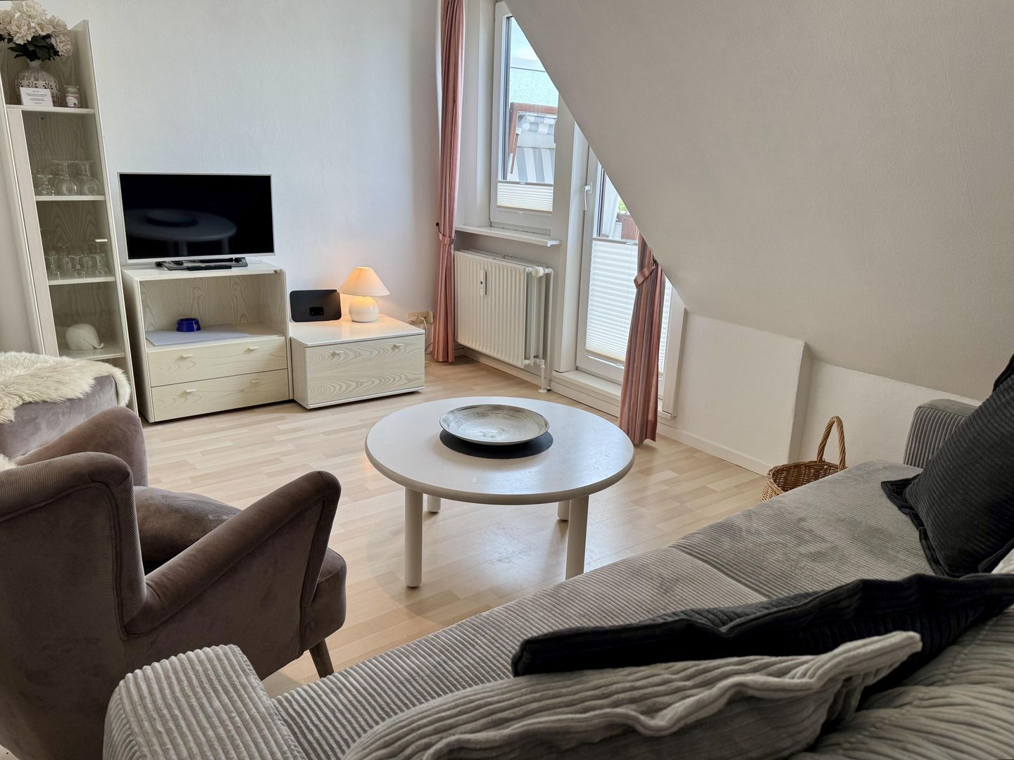 A modern living room with a sofa, a small round table, a TV on a stand, a bookshelf, and a window with curtains.
