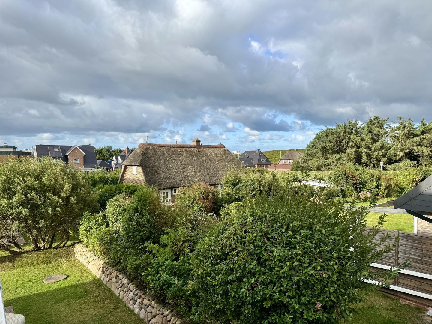 A thatched-roof house surrounded by dense greenery, with a stone wall and a grassy lawn.