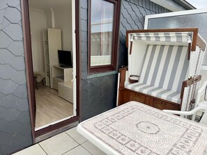 A room with a tiled floor, a small table with a patterned tablecloth, a striped chair, a television, and a cabinet. - SyltRuhe - 2-room apartment for 4 people with a balcony, parking space, and Wi-Fi. Pet (Sylt)