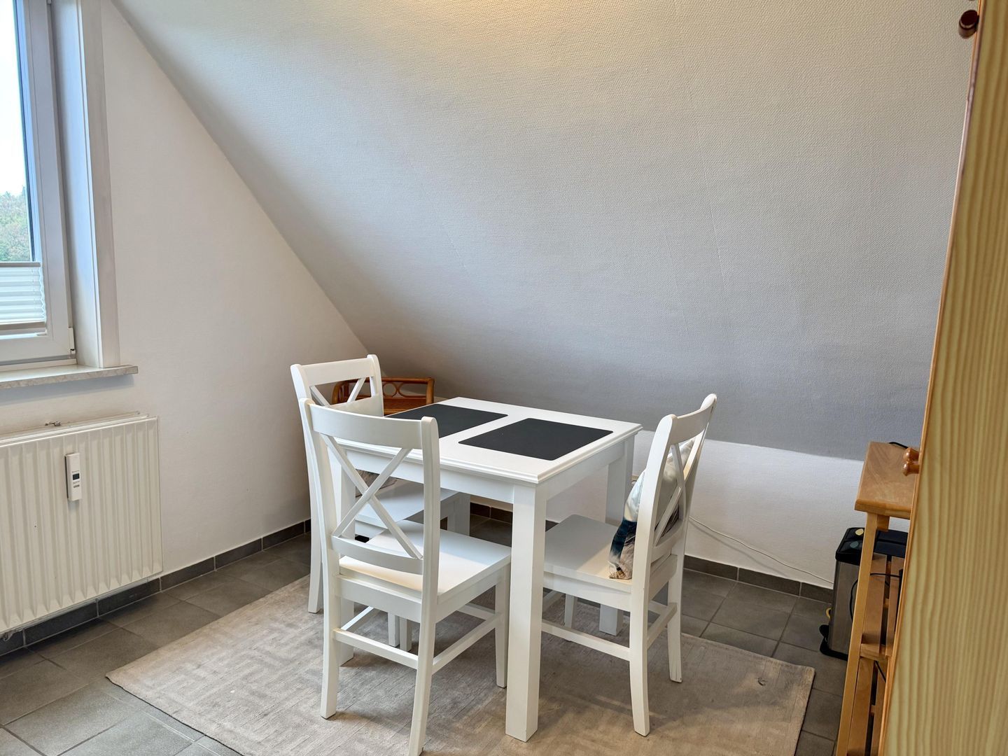 Une pièce avec une table et des chaises blanches, un radiateur, une fenêtre avec des stores et un coin avec une chaise en bois.