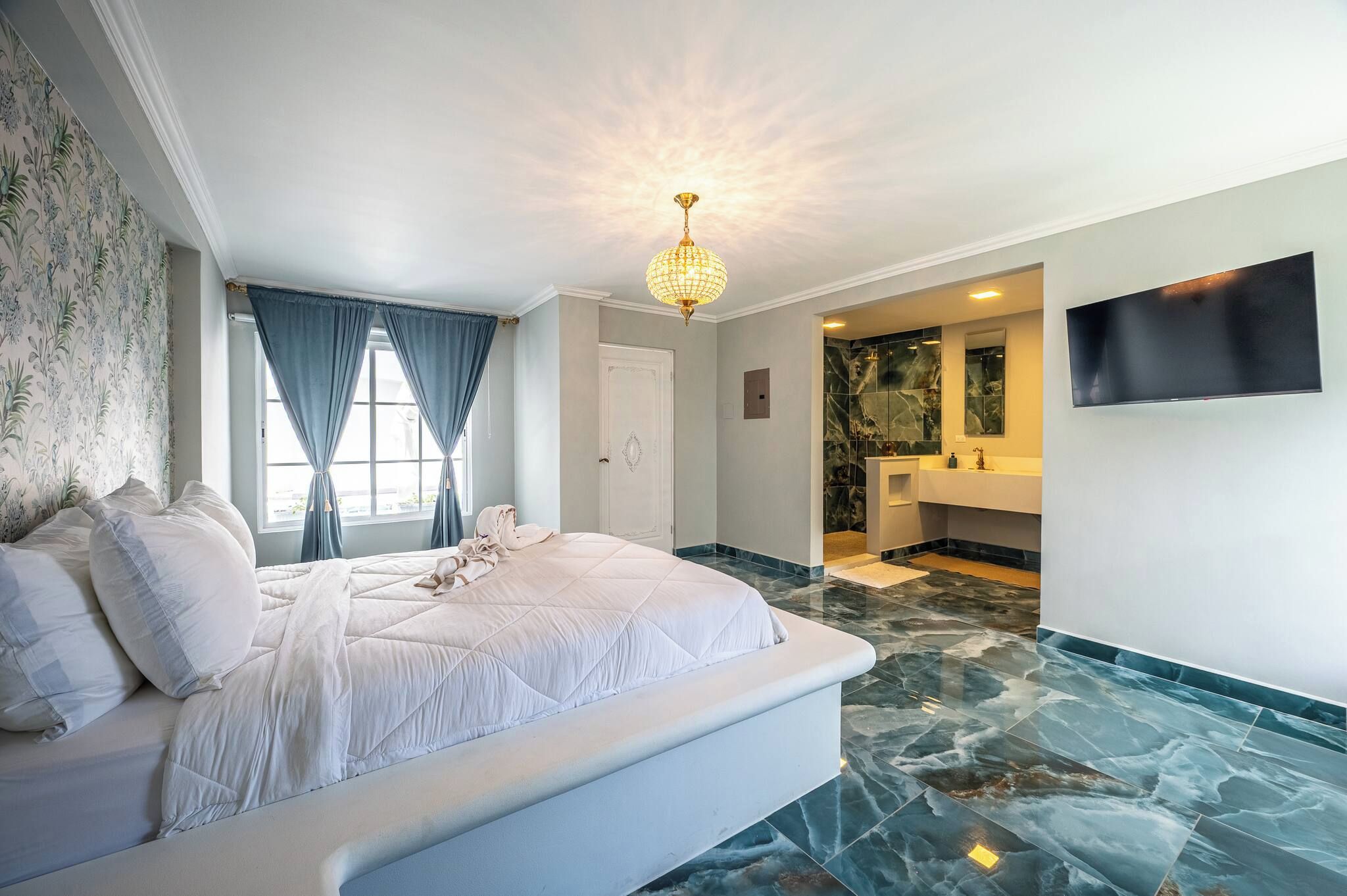 Une chambre à coucher avec un lit, un lustre, une salle de bain avec un miroir et un lavabo, et un téléviseur fixé au mur.