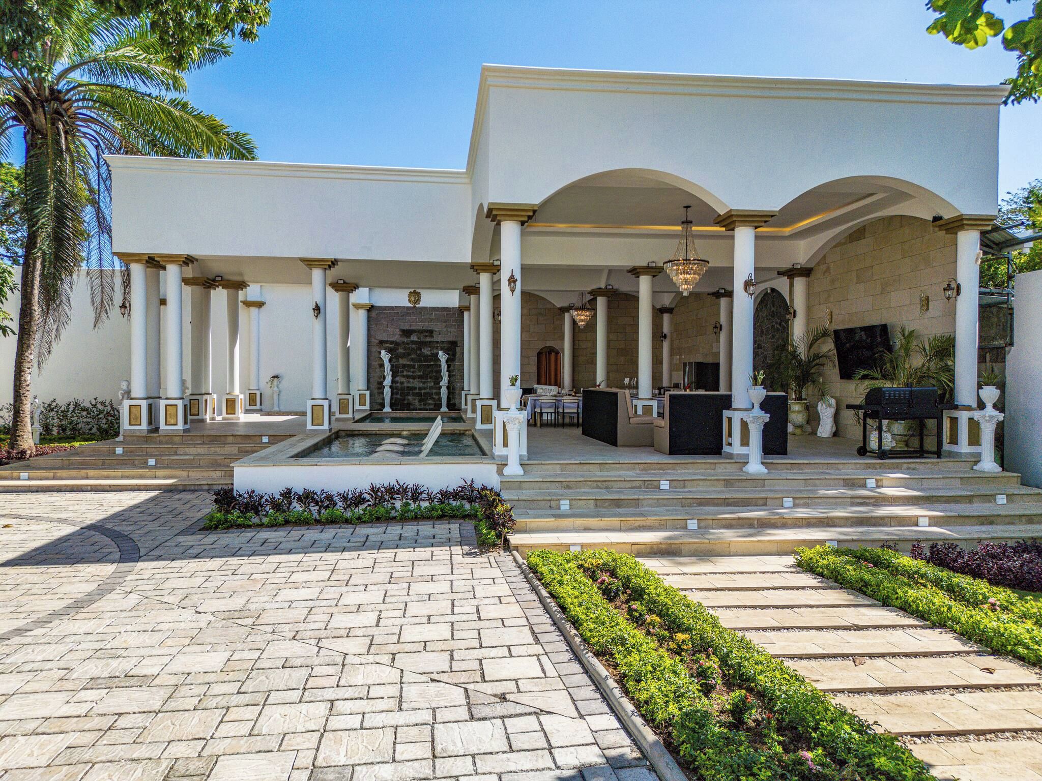 Un edificio con acceso cubierto, columnas, una fuente y un jardín ajardinado.