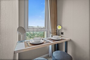A modern hotel room with a dining area, a window with a view, a bedside table with a lamp, and a chair. - Eslin by Kozystay - Cilandak (South Jakarta)